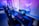 A row of modern gaming setups is arranged neatly along a window-lined wall. Each station includes a high-resolution monitor displaying vivid graphics, a mechanical keyboard with colored lighting, a gaming mouse, and an ergonomic chair. The room is illuminated with ambient blue and purple lights, creating a sleek and futuristic atmosphere.