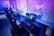 A row of modern gaming setups is arranged neatly along a window-lined wall. Each station includes a high-resolution monitor displaying vivid graphics, a mechanical keyboard with colored lighting, a gaming mouse, and an ergonomic chair. The room is illuminated with ambient blue and purple lights, creating a sleek and futuristic atmosphere.
