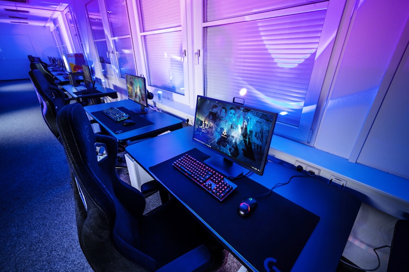 A row of modern gaming setups is arranged neatly along a window-lined wall. Each station includes a high-resolution monitor displaying vivid graphics, a mechanical keyboard with colored lighting, a gaming mouse, and an ergonomic chair. The room is illuminated with ambient blue and purple lights, creating a sleek and futuristic atmosphere.