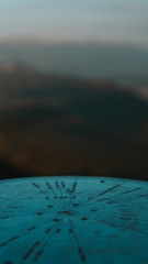 A metal compass or directional dial is mounted atop a surface, with various distances and directions inscribed on it. The background is an out-of-focus landscape, possibly featuring mountains or hills under a soft sky.