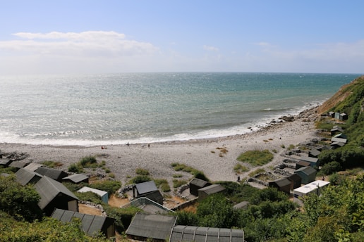 A coastal scene with a pebble beach stretching along the shoreline. Small cabins or huts are scattered across the hillside, surrounded by green vegetation. The sea looks slightly choppy with small waves, and a few people are seen walking along the beach. The sky above is partly cloudy with patches of blue.