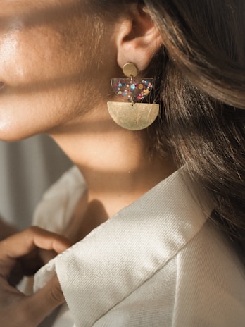 A close-up of a woman's ear, adorned with an artistic earring consisting of a golden semi-circle and a transparent piece with colorful confetti. Her hair is brown and flows naturally, while she wears a white textured shirt. Soft sunlight casts gentle shadows, adding an elegant touch.
