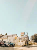 A small town setting with vintage buildings and vehicles. An old, rustic gas station sign reads 'OPAL' and stands prominently in the scene. The buildings have a weathered look, suggesting they've been here for a long time. Pickup trucks are parked nearby, and trees rise in the background under a clear sky.