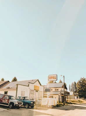 A small town setting with vintage buildings and vehicles. An old, rustic gas station sign reads 'OPAL' and stands prominently in the scene. The buildings have a weathered look, suggesting they've been here for a long time. Pickup trucks are parked nearby, and trees rise in the background under a clear sky.