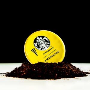 A yellow Starbucks Veranda Blend Americano lid is partially embedded in a pile of ground coffee against a stark black background. The contrast highlights the brand's logo and text on the lid.