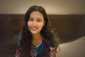 A woman with long, dark hair smiles warmly. She wears a colorful, patterned shirt and is seated indoors against a neutral background. The lighting is soft, creating a cozy and intimate atmosphere.