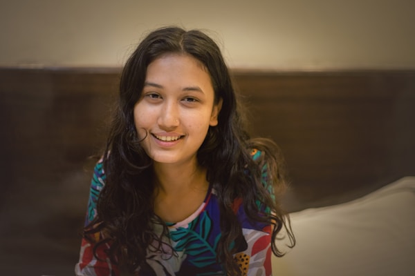 A woman with long, dark hair smiles warmly. She wears a colorful, patterned shirt and is seated indoors against a neutral background. The lighting is soft, creating a cozy and intimate atmosphere.