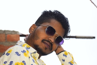 A man with a short beard wearing sunglasses and a floral shirt with yellow and blue patterns is leaning against a wall made of red bricks. His hair is styled neatly, and the background appears to be an outdoor setting.