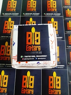 A set of promotional materials featuring the logo and branding for 'Big Eaters'. The central focal point is a box with the logo displayed prominently in orange and yellow against a dark background. Surrounding the box are several flyers or cards with the same logo and contact information repeated, including phone numbers and social media handles.