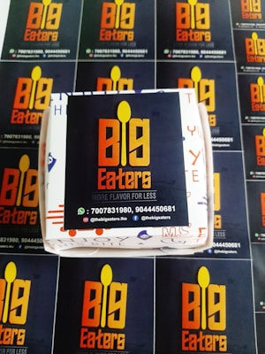 A set of promotional materials featuring the logo and branding for 'Big Eaters'. The central focal point is a box with the logo displayed prominently in orange and yellow against a dark background. Surrounding the box are several flyers or cards with the same logo and contact information repeated, including phone numbers and social media handles.