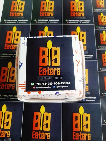 A set of promotional materials featuring the logo and branding for 'Big Eaters'. The central focal point is a box with the logo displayed prominently in orange and yellow against a dark background. Surrounding the box are several flyers or cards with the same logo and contact information repeated, including phone numbers and social media handles.