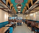 An elegantly designed restaurant interior with intricately patterned ceilings and ornate light fixtures. The walls are decorated with artistic murals, and the floor boasts a geometric tile pattern. The seating includes plush, teal-colored sofa benches and dark wooden tables, with gold-toned cups set neatly on the tables.