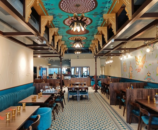 An elegantly designed restaurant interior with intricately patterned ceilings and ornate light fixtures. The walls are decorated with artistic murals, and the floor boasts a geometric tile pattern. The seating includes plush, teal-colored sofa benches and dark wooden tables, with gold-toned cups set neatly on the tables.