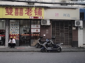 A small storefront with Chinese characters on the sign, featuring a display of food items in the window. Two mopeds are parked outside on the street, and a man in a white shirt is sitting on the curb using a phone. The adjacent store has a closed gate and air conditioning units above.