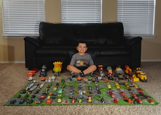 A smiling child sits on a carpet in front of a black sofa, surrounded by a collection of toy cars and trucks arranged neatly on a play mat. The play mat has roads and green areas depicted, enhancing the scene with a pretend city layout.