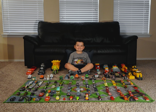 A smiling child sits on a carpet in front of a black sofa, surrounded by a collection of toy cars and trucks arranged neatly on a play mat. The play mat has roads and green areas depicted, enhancing the scene with a pretend city layout.