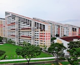 A group of modern residential buildings featuring a unique slanted design, surrounded by a lush green lawn and several trees. The buildings have multiple floors with a light color scheme, including white and pastel shades. The area around the buildings is well-maintained with pathways and some small decorative elements near the grass.