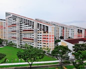 A group of modern residential buildings featuring a unique slanted design, surrounded by a lush green lawn and several trees. The buildings have multiple floors with a light color scheme, including white and pastel shades. The area around the buildings is well-maintained with pathways and some small decorative elements near the grass.