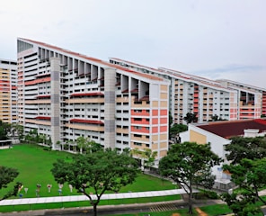 A group of modern residential buildings featuring a unique slanted design, surrounded by a lush green lawn and several trees. The buildings have multiple floors with a light color scheme, including white and pastel shades. The area around the buildings is well-maintained with pathways and some small decorative elements near the grass.
