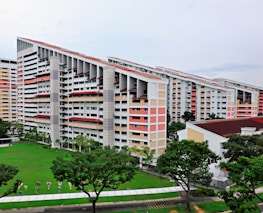 A group of modern residential buildings featuring a unique slanted design, surrounded by a lush green lawn and several trees. The buildings have multiple floors with a light color scheme, including white and pastel shades. The area around the buildings is well-maintained with pathways and some small decorative elements near the grass.