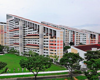 A group of modern residential buildings featuring a unique slanted design, surrounded by a lush green lawn and several trees. The buildings have multiple floors with a light color scheme, including white and pastel shades. The area around the buildings is well-maintained with pathways and some small decorative elements near the grass.
