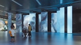 A modern interior space with large, digital display panels showing abstract patterns and text in blue tones. People, including a person with a stroller and a child, are observing the displays. The ceiling has a grid design with lights, and the floor is reflective, enhancing the ambient lighting.