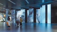 A modern interior space with large, digital display panels showing abstract patterns and text in blue tones. People, including a person with a stroller and a child, are observing the displays. The ceiling has a grid design with lights, and the floor is reflective, enhancing the ambient lighting.