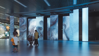 A modern interior space with large, digital display panels showing abstract patterns and text in blue tones. People, including a person with a stroller and a child, are observing the displays. The ceiling has a grid design with lights, and the floor is reflective, enhancing the ambient lighting.
