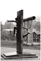 A sculpture of a human figure appears to be emerging through a vertical slab. The scene is outdoors with an overcast sky. In the background, there are portions of painted wall segments and a metal railing.