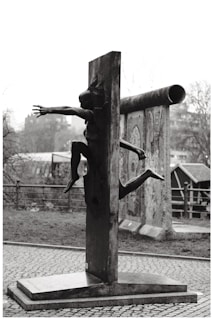 A sculpture of a human figure appears to be emerging through a vertical slab. The scene is outdoors with an overcast sky. In the background, there are portions of painted wall segments and a metal railing.