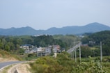 A serene rural landscape featuring a small village nestled among lush green hills. Low-rise houses with colorful roofs are scattered along a winding road that stretches into the distance. Towering trees and dense vegetation surround the area, and gentle hills rise in the background under a calm sky.
