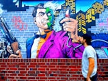 Graffiti art depicting a cartoonish villain character with a dual-colored face, wearing a vibrant purple and orange suit. The character is holding a large shotgun and a spray can emitting smoke, set against an urban backdrop featuring a city skyline and brick wall. A person in a baseball cap and casual attire gazes at the mural.