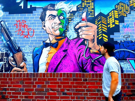 Graffiti art depicting a cartoonish villain character with a dual-colored face, wearing a vibrant purple and orange suit. The character is holding a large shotgun and a spray can emitting smoke, set against an urban backdrop featuring a city skyline and brick wall. A person in a baseball cap and casual attire gazes at the mural.