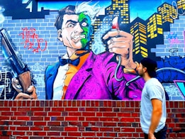 Graffiti art depicting a cartoonish villain character with a dual-colored face, wearing a vibrant purple and orange suit. The character is holding a large shotgun and a spray can emitting smoke, set against an urban backdrop featuring a city skyline and brick wall. A person in a baseball cap and casual attire gazes at the mural.