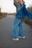 A person stands on a road, wearing a plaid top and flared blue jeans, with a denim jacket casually draped over the shoulders. The scene is set outdoors, with tall grass lining the road and a blurred background suggesting a rural environment.