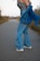A person stands on a road, wearing a plaid top and flared blue jeans, with a denim jacket casually draped over the shoulders. The scene is set outdoors, with tall grass lining the road and a blurred background suggesting a rural environment.