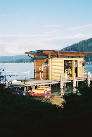 A person wearing swimwear is standing on a wooden dock next to a small wooden building by the water. The building is labeled with a sign that says 'SEESport' and has various equipment hanging inside, including life jackets. There is a mountainous landscape in the background and several brightly colored inflatable rafts docked in the water in the foreground.