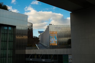 A modern architectural structure with metallic and glass surfaces is seen under a partly cloudy sky. A colorful banner with the word 'Solidarity' is hung on one of the buildings. The scene is framed by concrete structures, and there is some greenery visible.