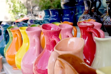 A collection of colorful ceramic pitchers arranged in rows, featuring a variety of bright hues including pink, red, yellow, and blue. The ceramics are glossy and reflect light, creating a vibrant display. The background shows other pottery pieces with intricate patterns.