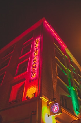 A multistory building is illuminated with colorful neon lights. The sign vertically spells out 'HOTEL DE FRANCE', using vibrant red and yellow hues. The glow from the signage reflects onto the building’s façade, giving it a lively and warm appearance. A small circular sign at street level reads 'INTER-HOTEL'. The night sky and street lighting add to an atmospheric urban ambiance.