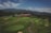 A sprawling golf course with vibrant green fairways and sand bunkers is set against a backdrop of rolling hills and mountains. A clubhouse with a red roof and multiple gables is centrally located, surrounded by landscaped pathways and bushes. The sky above is partly cloudy.
