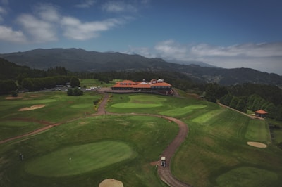 A sprawling golf course with vibrant green fairways and sand bunkers is set against a backdrop of rolling hills and mountains. A clubhouse with a red roof and multiple gables is centrally located, surrounded by landscaped pathways and bushes. The sky above is partly cloudy.