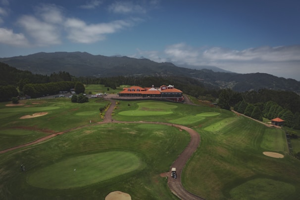 A sprawling golf course with vibrant green fairways and sand bunkers is set against a backdrop of rolling hills and mountains. A clubhouse with a red roof and multiple gables is centrally located, surrounded by landscaped pathways and bushes. The sky above is partly cloudy.