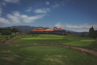 A lush green landscape features a golf course with neatly manicured grass. A large building with a red roof and multiple windows is situated in the background, surrounded by distant hills. The sky is partly cloudy, creating a serene and peaceful atmosphere.