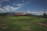 A lush green landscape features a golf course with neatly manicured grass. A large building with a red roof and multiple windows is situated in the background, surrounded by distant hills. The sky is partly cloudy, creating a serene and peaceful atmosphere.