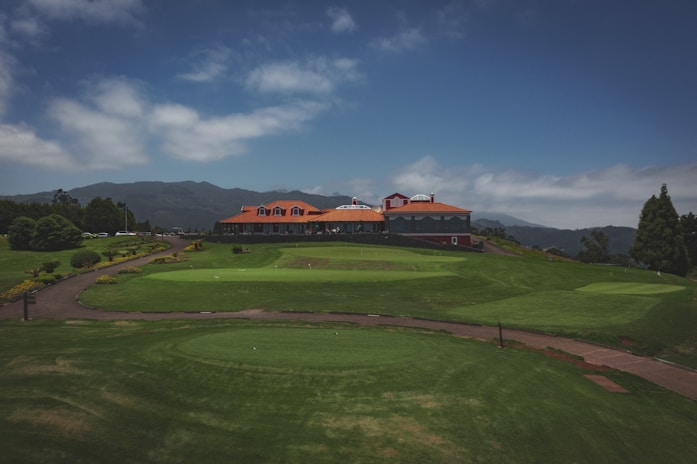 A lush green landscape features a golf course with neatly manicured grass. A large building with a red roof and multiple windows is situated in the background, surrounded by distant hills. The sky is partly cloudy, creating a serene and peaceful atmosphere.
