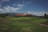 A lush green landscape features a golf course with neatly manicured grass. A large building with a red roof and multiple windows is situated in the background, surrounded by distant hills. The sky is partly cloudy, creating a serene and peaceful atmosphere.