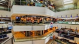 A multi-level shopping mall with numerous shops and stores visible. Bright, artificial lighting illuminates the interior, with glass railings surrounding the walkways. Various signs and advertisements are prominently displayed, and mannequins can be seen through the glass storefronts. Customers are walking in the thoroughfares.