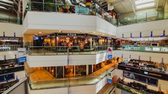 A multi-level shopping mall with numerous shops and stores visible. Bright, artificial lighting illuminates the interior, with glass railings surrounding the walkways. Various signs and advertisements are prominently displayed, and mannequins can be seen through the glass storefronts. Customers are walking in the thoroughfares.
