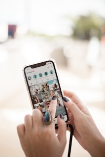 A person's hand is holding a smartphone with a social media application open, displaying a grid of photos and a username with the word 'JUNE' in bold letters. The nails are painted dark, and the person is wearing a large ring. The background is blurred, suggesting an outdoor setting.